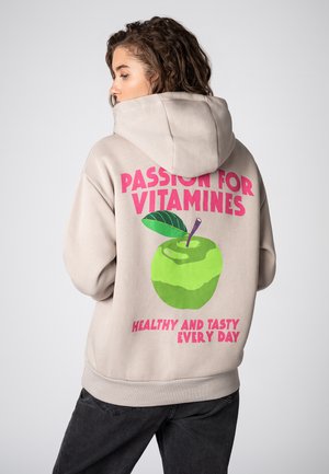 Beige Kapuzenpullover mit einem großen grünen Apfel-Graphic auf dem Rücken, der Text lautet "LEIDENSCHAFT FÜR VITAMINE" und "GESUND UND LECKER JEDEN TAG" in Pink.