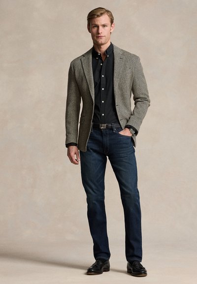 Man draagt een visgraat tweed blazer over een zwart overhemd met knopen, donkerblauwe jeans, zwarte riem en zwarte leren schoenen, staand met hand in zak.