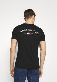 Camiseta de algodón negra con el texto "TOMMY HILFIGER" y un pequeño logo con una bandera en rojo, blanco y azul; de mangas cortas y cuello redondo.