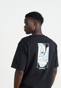 Black cotton t-shirt featuring a graphic print in blue and cream on the back with "new balance" text at the bottom. Round neckline, short sleeves.