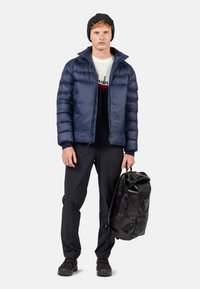 Giacca imbottita blu navy con colletto alto, berretto in maglia nero, maglione bianco con dettagli rossi, pantaloni scuri e uno zaino impermeabile nero.