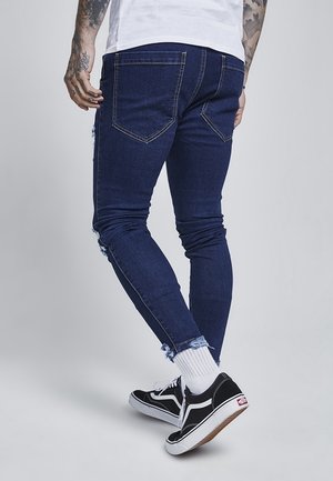 Mężczyzna ubrany w ciemnoniebieskie obcisłe dżinsy z postrzępionymi brzegami, białe skarpetki, czarne trampki i biały t-shirt, stojący na tle jednolitego tła.