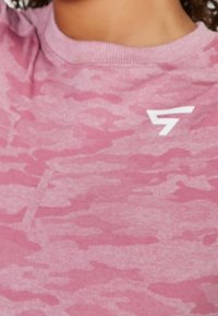 Gros plan d'une personne portant une chemise rose à motif camouflage avec un logo géométrique blanc sur le haut de la poitrine.