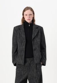 Črno-siva karirana blazer jakna z zobatimi reverji, dvema gumboma in rahlo ohlapnim krojem, v kombinaciji s črnim rebrastim topom z visokim ovratnikom.