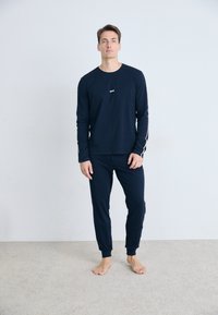 Mann som står barføtt på teppet, iført en marineblå skjorte med lange ermer og matchende joggebukser med hvite striper på ermene.