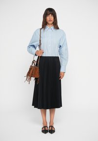 Chemise à rayures bleues et blanches avec des manches bouffantes, associée à une jupe plissée noire. Accessoirisée avec un sac à main à franges marron.