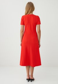 Een vrouw staat met haar rug naar de camera, gekleed in een kniehoge rode jurk en zwarte sandalen, voor een neutrale, minimalistische achtergrond.