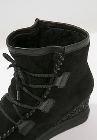 Bottine compensée en daim noir avec lacets à l’avant, bordure en cuir et détail texturé au talon.