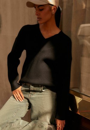 Svart ribbad tröja med V-ringning, bärs med ljusblå högmidjade jeans. Guldörhängen och en beige keps fulländar outfiten.
