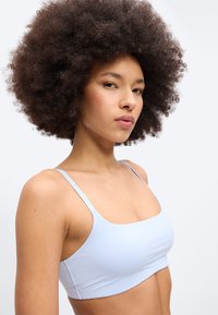 Νεαρή γυναίκα με πυκνά σγουρά μαλλιά που φοράει ένα ανοιχτό γαλάζιο τοπάκι με λεπτές τιράντες, πάνω σε απλό φόντο.