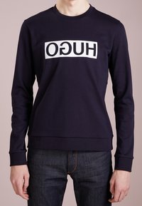 Marinblå sweatshirt med vit text "HUGO" tryckt horisontellt. Har rund halsringning och ribbstickade muddar och fåll. Tyget är mjukt och elastiskt.