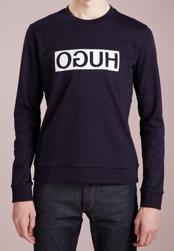 Marinblå sweatshirt med vit text "HUGO" tryckt horisontellt. Har rund halsringning och ribbstickade muddar och fåll. Tyget är mjukt och elastiskt.