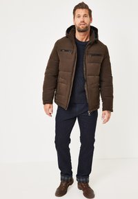 Braune, gepolsterte Jacke mit Kapuze, ausgestattet mit zwei Reißverschluss-Brusttaschen. Getragen über einem marineblauen Pullover und dunklen Jeans mit karierter Saum.