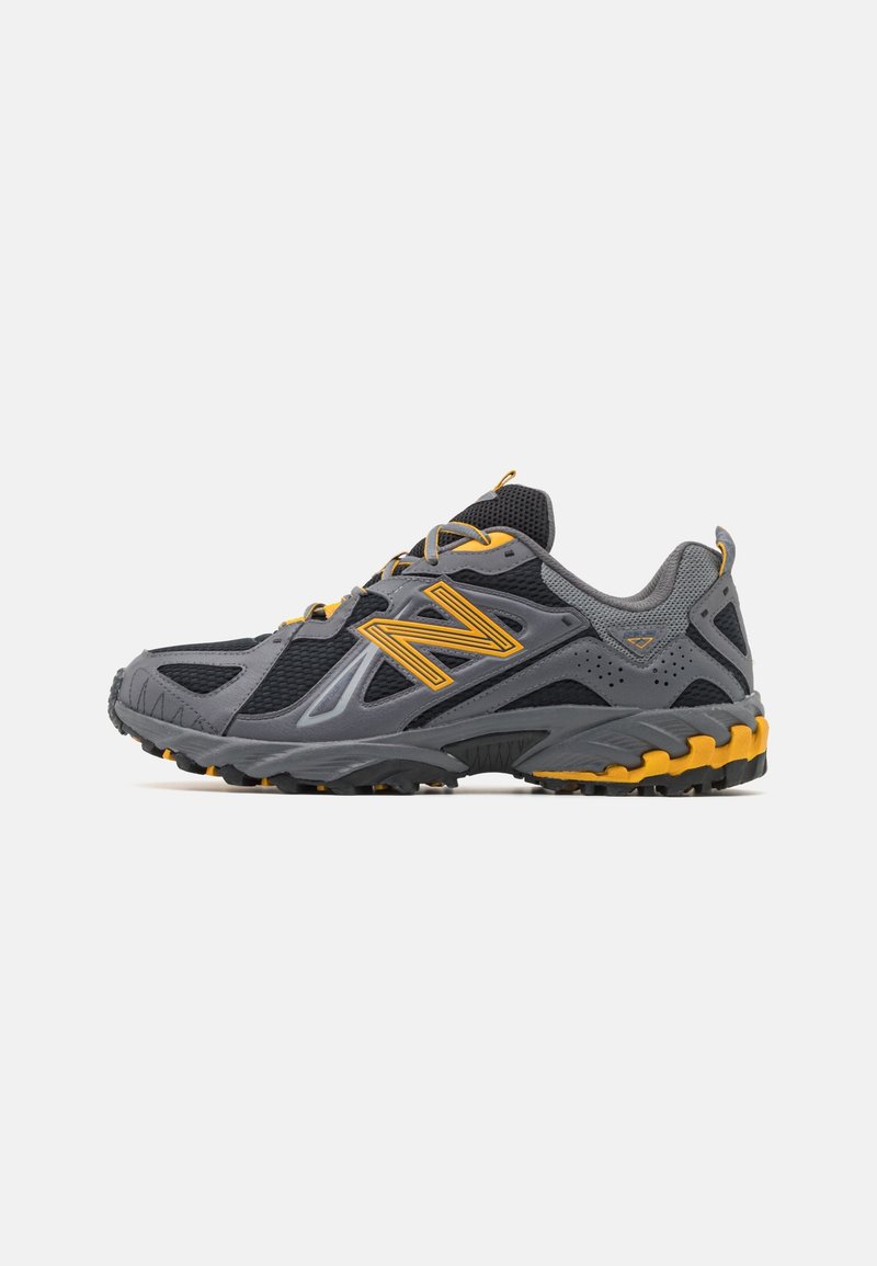 Chaussure de trail running grise et noire avec des touches jaunes, semelle épaisse à crampons, panneaux en maille et grand logo jaune « N » sur le côté.