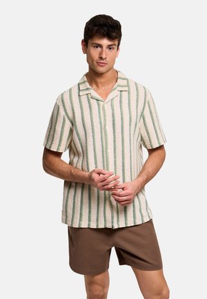 Hombre con camisa de manga corta color crema con rayas verticales verdes y pantalones cortos marrones, de pie con las manos entrelazadas a la altura de la cintura.