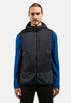 Schwarze ärmellose Weste mit Kapuze, mit Reißverschluss vorne, grauem strukturiertem unteren Teil und Logo auf der Brust. Getragen über einem blauen Langarmshirt.