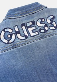 Kurtka dżinsowa z granatowym haftowanym napisem "GUESS" na górnej części pleców, z klasycznym kołnierzykiem i kontrastującymi detalami przeszycia.