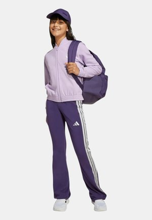 Lächendes Mädchen trägt einen lila Trainingsanzug, eine Kappe, weiße Turnschuhe und trägt einen großen lila Rucksack über einer Schulter.