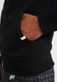 Una mano di carnagione scura parzialmente dentro la tasca anteriore di una felpa con zip nera, indossata con pantaloni a quadri blu e un ciondolo di perline attaccato alla cintura.
