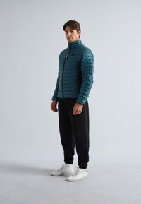 Doudoune bleu canard avec des coutures horizontales, col haut et poches latérales noires, portée avec un pantalon noir et des baskets blanches.