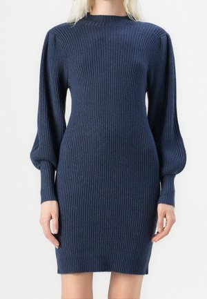 Femme portant une robe en maille côtelée bleu marine avec de longues manches bouffantes et un col montant.