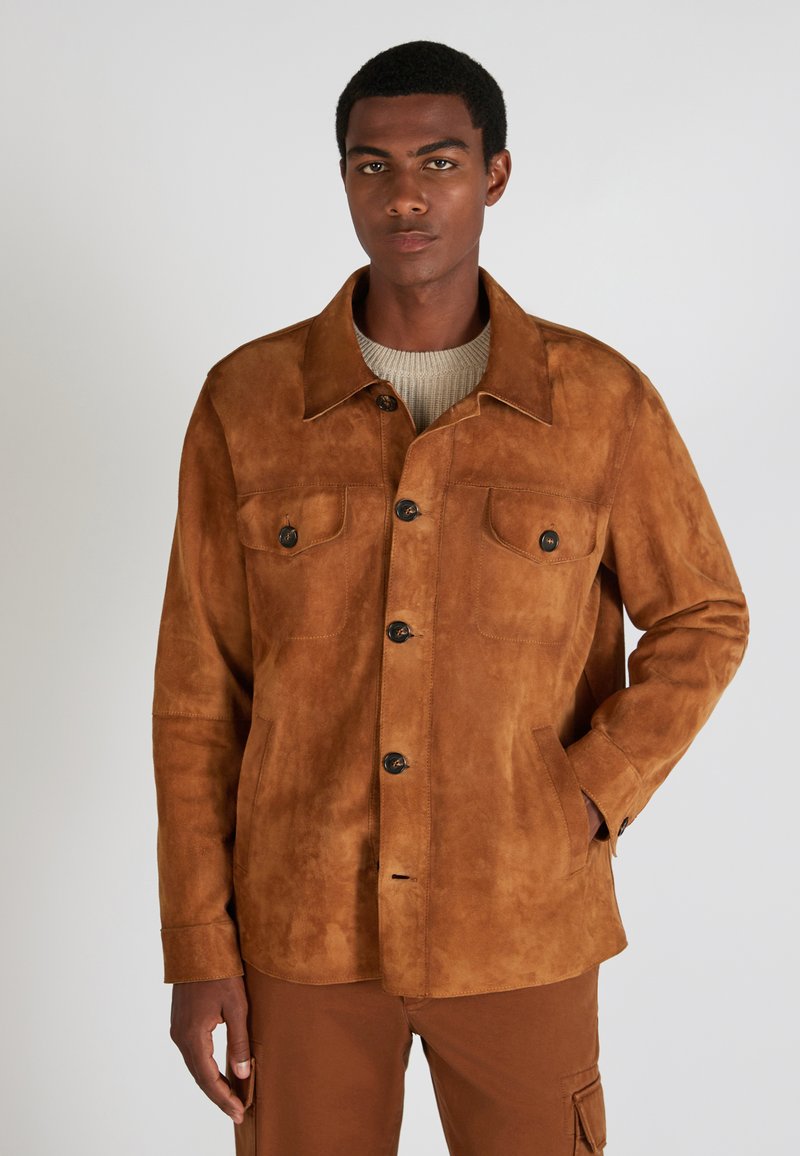 Braune Wildlederjacke mit zwei Brusttaschen, schwarzen Knöpfen und einem geraden Kragen, kombiniert mit einem hellen Pullover darunter.