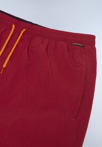 Shorts légers rouges avec taille élastique et cordon orange, dotés de poches latérales et d'une surface texturée.