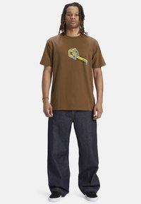 Brown cotton t-shirt featuring a tape measure graphic in yellow and gray. Paired with loose-fitting dark denim jeans and black sneakers.