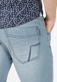 Light blue denim jeans featuring a curved back pocket with contrasting stitching and a subtle wear texture. Pair of navy patterned shirt peeking out.