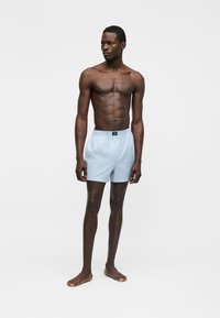 Bărbat slab cu piele închisă la culoare, purtând șorturi drepte cu dungi albastre deschise și albe, stând desculț în fața unui fundal alb simplu.