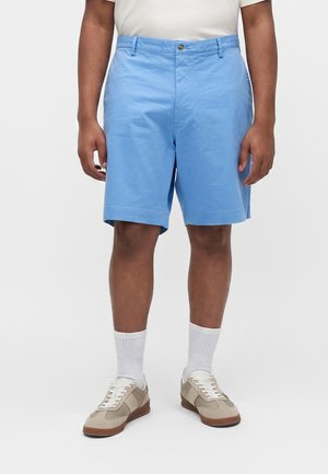 Person iført lyseblå shorts, hvide crew-sokker, beige og hvide sneakers samt en hvid skjorte, stående foran en ensfarvet baggrund.