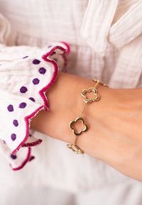 Bracciale d'oro con collegamenti a forma di quadrifoglio e accenti circolari, abbinato a una blusa bianca testurizzata con pois viola e rifiniture rosa.