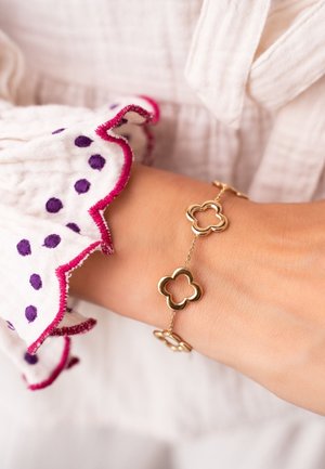 Bracciale d'oro con collegamenti a forma di quadrifoglio e accenti circolari, abbinato a una blusa bianca testurizzata con pois viola e rifiniture rosa.