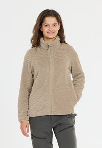 Beige fleece jack met golvende textuur, hoge kraag en een volledige ritssluiting. Draag het over donkergrijze broek met ritszakken.