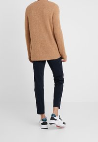 Maglione marrone lavorato a maglia con finitura strutturata, abbinato a pantaloni slim fit blu scuro e sneakers bianche con dettagli neri e turchesi.