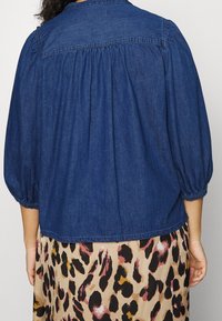 Donna che indossa una blusa di denim blu con maniche a sbuffo a tre quarti e una gonna con un motivo a stampa leopardata beige e multicolore.