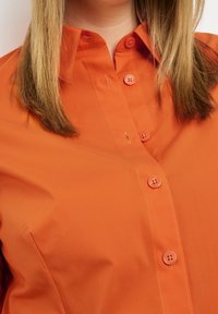 Camicia arancione con bottoni, caratterizzata da una texture liscia, colletto classico e sei bottoni coordinati sul davanti, con dettagli di cucitura discreti.