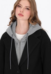 Črn plašč z teksturiranim zaključkom, položen čez sivi zip-up kapuco z vrvicami. Bel osnovni sloj je viden spodaj.