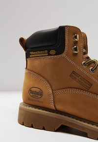 Bota de cuero impermeable marrón con un cuello acolchado negro, ojales dorados y una suela de goma resistente. Presenta una etiqueta que dice "repelente al agua".