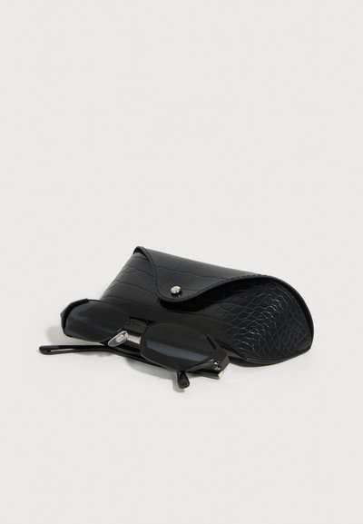 Μαύρα οκταγωνικά γυαλιά ηλίου που ακούν δίπλα σε μια μαύρη υφασμάτινη σκληρή θήκη με κλείσιμο με κουμπί σε ανοιχτή επιφάνεια.