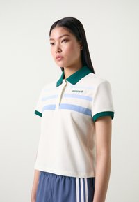 Camisa polo em algodão creme com colarinho e detalhes nas mangas em verde. Apresenta riscas horizontais em azul claro e um logótipo bordado. Design cropped.