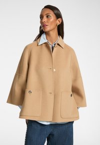 Beige ullblandad kappa med en bred silhuett, knäppt stängning, två stora framfickor och en spetsig krage. Bärs över en ljusblå skjorta.