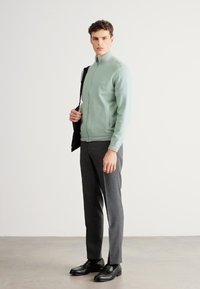 Ανοιχτό πράσινο τζάκετ με φερμουάρ, γκρι παντελόνι κατά παραγγελία και μαύρα παπούτσια· σε όρθια στάση, με μια μαύρη τσάντα περασμένη από τον έναν ώμο.