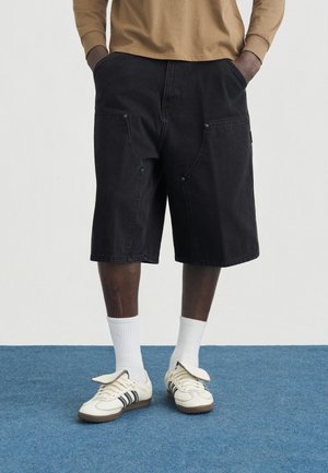 Persona che indossa pantaloncini neri fino al ginocchio, calzini bianchi e scarpe da ginnastica crema con strisce nere, in piedi su un tappeto blu con le mani in tasca.