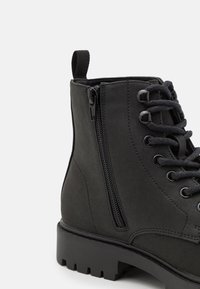 Botín negro hasta el tobillo con material de ante suave, parte delantera con cordones, cremallera lateral y suela de goma texturizada con un diseño de taco profundo.