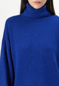 Μπλε πουλόβερ με ζιβάγκο, φτιαγμένο από μαλακό, υφασμένο νήμα. Διαθέτει ψηλό λαιμό και ρίγες στους ώμους για άνετη εφαρμογή.