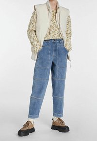 Béžová péřová vesta přes barevně pletený svetr, doplněná světle modrými džíny s vysokým pasem a hnědými platformovými botami na šněrování.