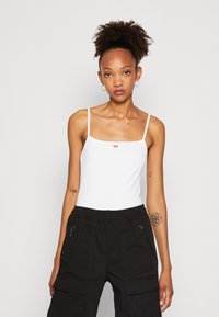 Hvit tettsittende tank topp med tynne stropper, med en liten rød logo. Kombinert med svarte cargo bukser med sidelommer med glidelås og elastisk midje.