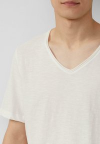 V-hals T-shirt gemaakt van lichtgewicht, zachte stof. Off-white kleur met een subtiele slub textuur en opgerolde mouwen, wat een ontspannen pasvorm benadrukt.