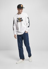Hvit langermet skjorte med svart "TUNE SQUAD" grafikk, blå jeans og hvite sneakers; iført en svart caps med en fargerik logo.